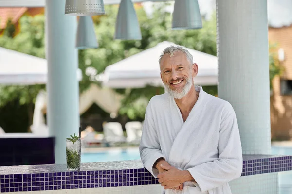 Fröhlicher reifer Mann in weißer Robe, der an der Poolbar neben Glas mit Mojito-Cocktail steht, Rückzug — Stockfoto