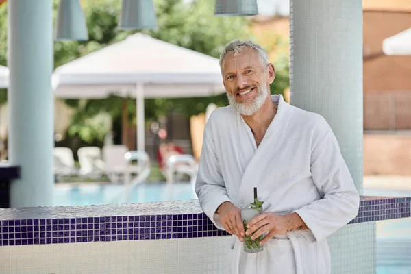 Positive mature man in white robe holding glass with mojito cocktail at poolside, wellness retreat — Stock Photo