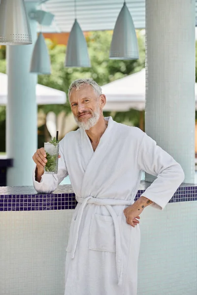 Homem maduro positivo em roupão branco segurando refrescante mojito cocktail na beira da piscina, retiro — Fotografia de Stock