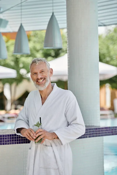 Middle aged man in white robe smiling and holding mojito cocktail at poolside, wellness retreat — Stock Photo