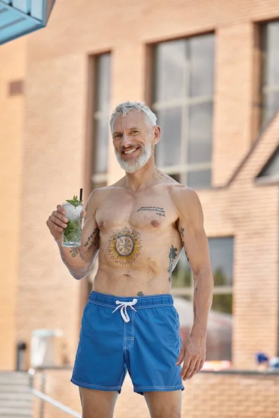 Tattooed middle aged man in shorts smiling and holding mojito cocktail on vacation, wellness retreat — Stock Photo