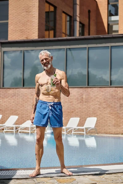 Татуированный мужчина средних лет в шортах улыбается и держит коктейль мохито у бассейна, оздоровительный отдых — стоковое фото