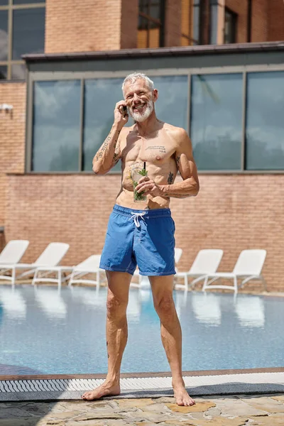 Glücklicher reifer Mann in kurzen Hosen, der mit dem Smartphone spricht und am Pool einen Cocktail hält, Wellness-Rückzugsort — Stockfoto