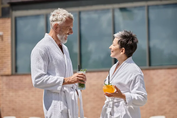 Felice coppia matura in accappatoi bianchi che tengono cocktail a bordo piscina, ritiro benessere in resort — Foto stock