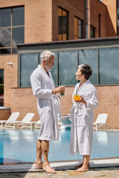 Cheerful middle aged couple in white robes holding cocktails at poolside, wellness retreat in resort — Stock Photo