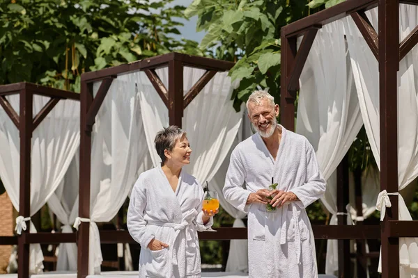 Cheerful middle aged couple in white robes holding cocktails near pavilion, wellness retreat — Stock Photo