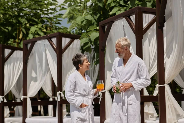 Happy mature couple in white robes holding cocktails and talking near pavilion, wellness retreat — Stock Photo