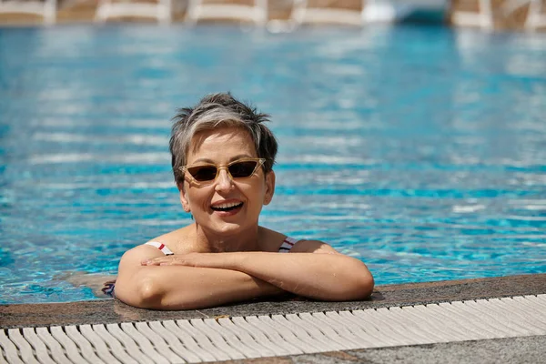Веселая женщина средних лет в солнцезащитных очках и купальниках внутри бассейна, оздоровительный отдых — стоковое фото