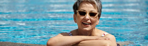 Cheerful middle aged woman in sunglasses and swimsuit inside of swimming pool, retreat, banner — Stock Photo