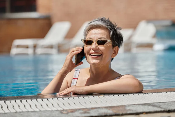Glückliche reife Frau mit Sonnenbrille, die im Inneren des Swimmingpools mit dem Smartphone spricht, Wellness-Rückzugsort — Stockfoto