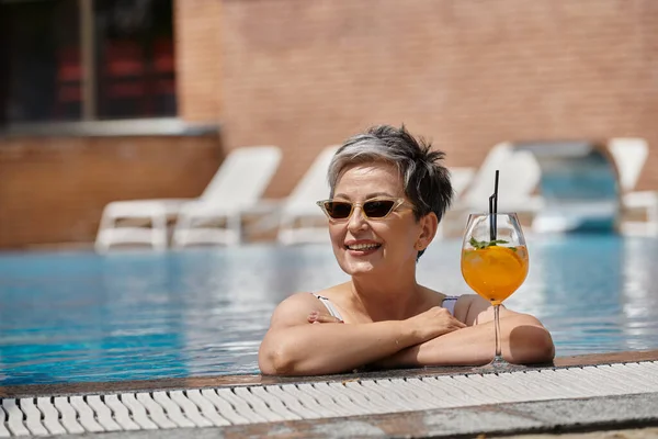 Fröhliche Frau mittleren Alters mit Sonnenbrille im Schwimmbad, am Pool mit Cocktail, Rückzug — Stockfoto