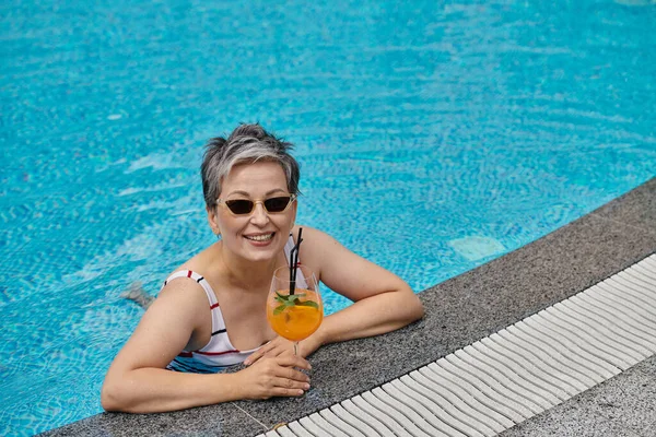Happy middle aged woman in sunglasses swimming in pool with blue water and holding cocktail, retreat — Stock Photo