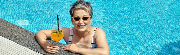 Happy mature woman in sunglasses swimming in pool with blue water and holding cocktail, banner — Stock Photo