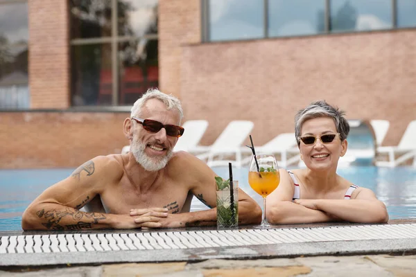 Feliz pareja madura en gafas de sol nadando en la piscina sonriendo cerca de cócteles, concepto de retiro de bienestar - foto de stock