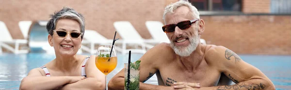 Feliz pareja madura en gafas de sol nadando en la piscina sonriendo cerca de cócteles, retiro de bienestar, pancarta - foto de stock