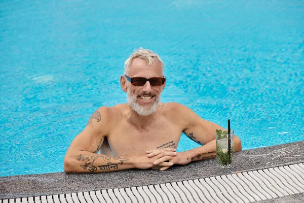 Без рубашки и татуировки мужчина средних лет в солнцезащитных очках внутри бассейна возле коктейля, отступление — стоковое фото