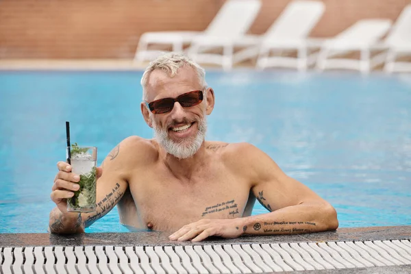 Torse nu et tatoué homme d'âge moyen dans des lunettes de soleil tenant cocktail et baignade dans la piscine, retraite — Photo de stock