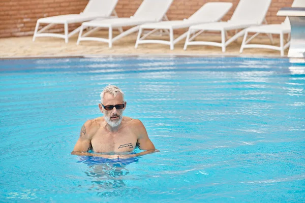 Без сорочки і татуйований чоловік середнього віку в сонцезахисних окулярах, який плаває в басейні з блакитною водою, відступає — стокове фото