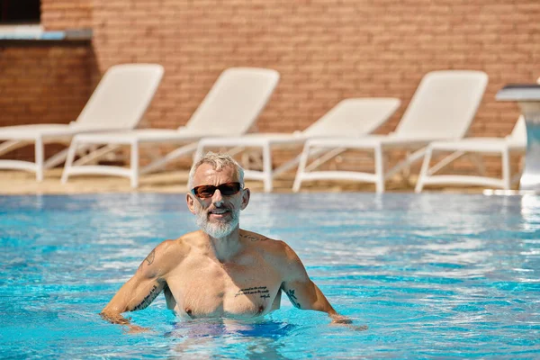 Hombre de mediana edad feliz y tatuado en gafas de sol nadando en la piscina con agua azul, retiro - foto de stock