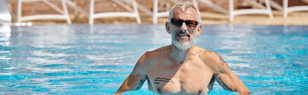 Счастливый и татуированный мужчина средних лет в солнцезащитных очках плавающий в бассейне с голубой водой, баннер — стоковое фото