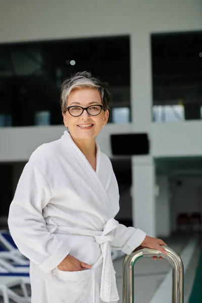 Happy middle aged woman in white robe and glasses smiling inside of indoor spa center, retreat — Stock Photo