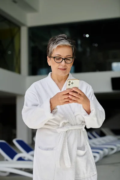 Mujer de mediana edad en bata blanca y gafas con teléfono inteligente dentro del centro de spa interior, retiro - foto de stock