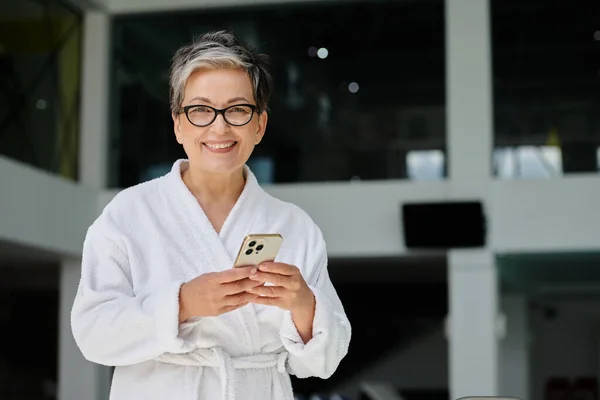 Happy mature woman in white robe and glasses using smartphone inside of indoor spa center, retreat — Stock Photo