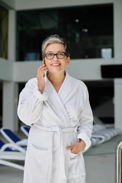 Happy mature woman in white robe and glasses talking on smartphone inside of spa center, retreat — Stock Photo
