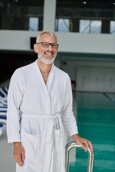Happy mature man in white robe and glasses smiling inside of indoor spa center, wellness retreat — Stock Photo