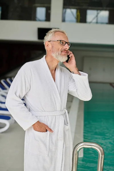 Tattooed mature man in white robe and glasses talking on smartphone inside of spa center, retreat — Stock Photo