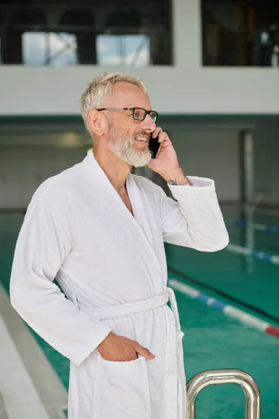 Homme mûr gai en robe blanche et lunettes parlant sur smartphone à l'intérieur du centre de spa, retraite — Photo de stock