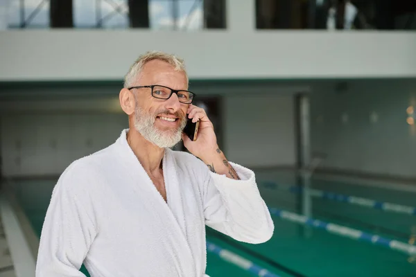 Homem maduro positivo em roupão branco e óculos falando no smartphone dentro do centro de spa, retiro — Fotografia de Stock