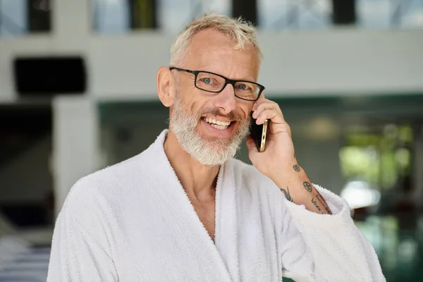 Hombre maduro positivo en bata blanca y gafas hablando en el teléfono inteligente dentro del centro de spa interior - foto de stock