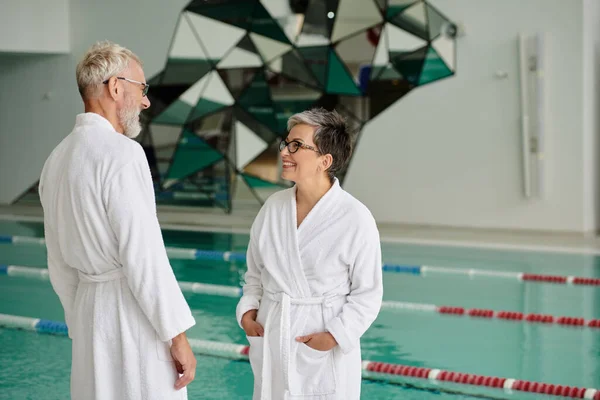 Cheerful middle aged couple in glasses and white robes chatting near indoor pool in spa center — Stock Photo