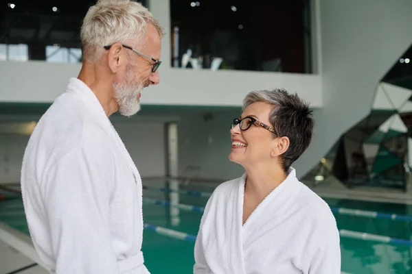 Joyful middle aged couple in glasses and white robes chatting near indoor pool in spa center — Stock Photo