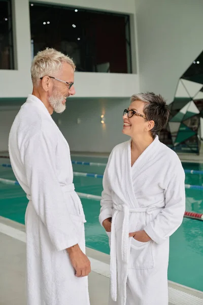 Alegre pareja de mediana edad en gafas y túnicas blancas de pie cerca de la piscina cubierta en el centro de spa - foto de stock