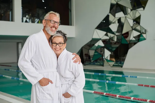 Feliz hombre de mediana edad en gafas y bata blanca abrazando a la esposa en el centro de spa, concepto de retiro de bienestar - foto de stock