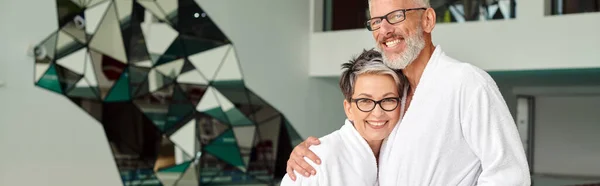 Happy middle aged man in glasses and white robe hugging wife in spa center, wellness retreat, banner — Stock Photo