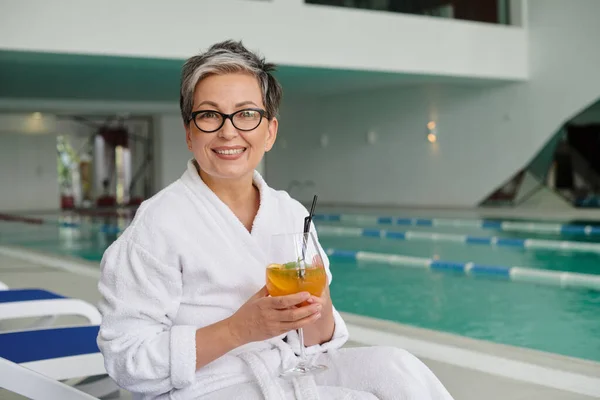 Spa center concept, happy mature woman in glasses and robe holding cocktail and sitting on lounger — Stock Photo