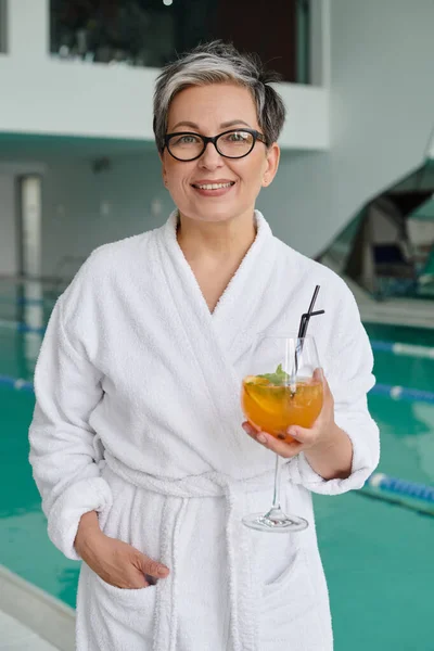 Concept spa et retraite, femme mûre heureuse en lunettes et peignoir tenant cocktail près de la piscine intérieure — Photo de stock
