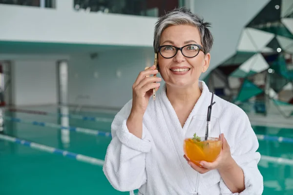 Rückzugskonzept, glückliche reife Frau in Brille und Robe, Cocktail in der Hand und Gespräche auf dem Smartphone — Stockfoto