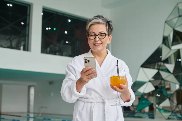 Concepto de bienestar y spa, mujer madura feliz en gafas celebración cóctel y el uso de teléfono inteligente - foto de stock