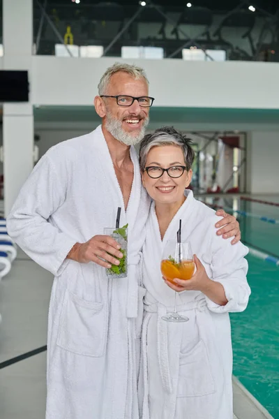 Happy middle aged couple in white robes holding glasses with cocktails in spa center near pool — Stock Photo