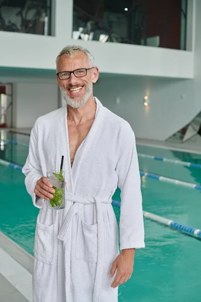 Concepto de retiro de bienestar, hombre maduro feliz en bata blanca celebración de cóctel cerca de la piscina en el centro de spa - foto de stock