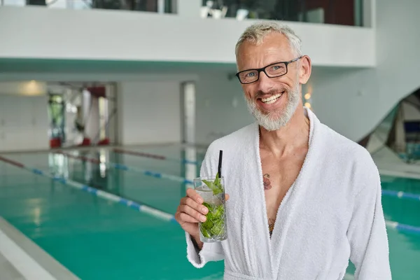 Веселый взрослый мужчина в белом халате держит коктейль возле бассейна в спа-центре — стоковое фото