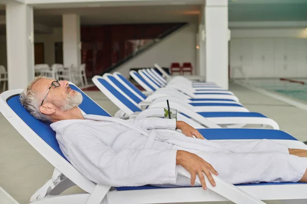 Оздоровительный ретрит концепция, зрелый мужчина в белом халате отдыхает на шезлонге возле коктейля в спа-центре — стоковое фото
