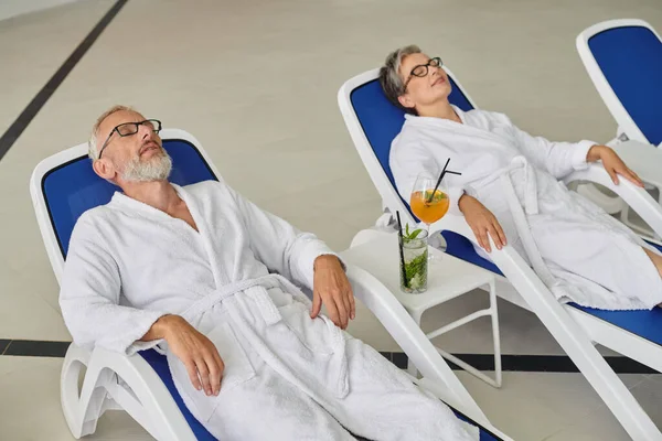 Rückzug-Konzept, reifes Paar in weißen Bademänteln auf Liegestühlen in der Nähe von Cocktails im Wellnesszentrum ausruhen — Stockfoto
