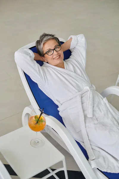 Concepto de retiro, mujer madura feliz en bata blanca descansando en la tumbona cerca de cóctel en el centro de spa - foto de stock