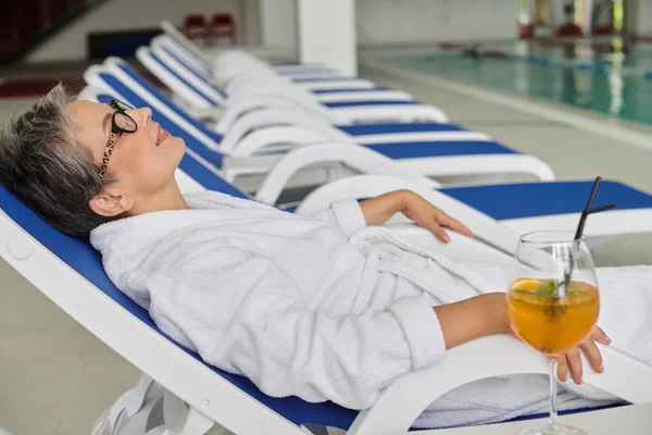 Spa retreat concept, pleased mature woman in white robe and glasses resting on lounger near cocktail — Stock Photo