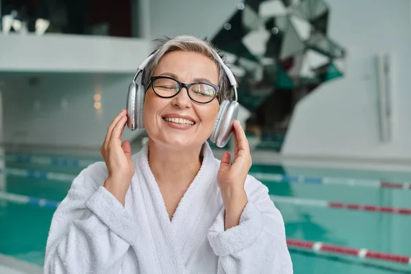 Happy middle aged woman in glasses and robe listening music in wireless headphones in spa center — Stock Photo